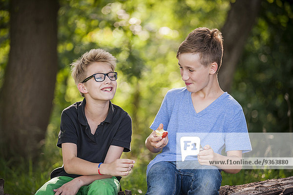 Zwei Jungen sitzen auf einem Baumstamm und essen einen Apfel.