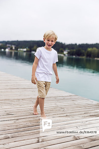 Porträt eines lächelnden kleinen Jungen  der auf einem Steg am See spazieren geht.