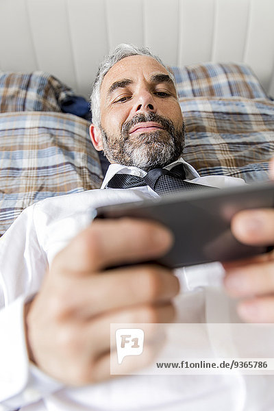Porträt eines auf dem Bett liegenden Geschäftsmannes mit Blick auf sein Smartphone