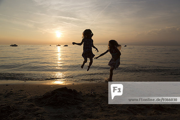Italy  Lake Garda  two girls jumping on beach at sunset