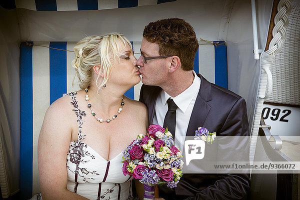 Porträt eines Brautpaares  das sich in einem Strandstuhl mit Kapuze küsst.