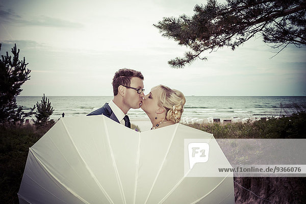 Brautpaar  das sich hinter einem Regenschirm vor dem Meer küsst.