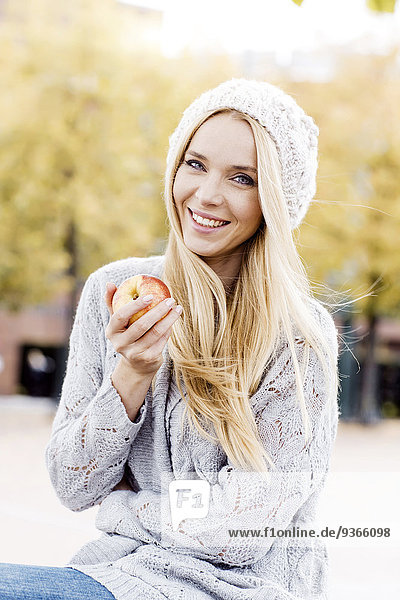 Porträt einer lächelnden jungen Frau  die einen Apfel mit Wollmütze und Strickjacke hält.