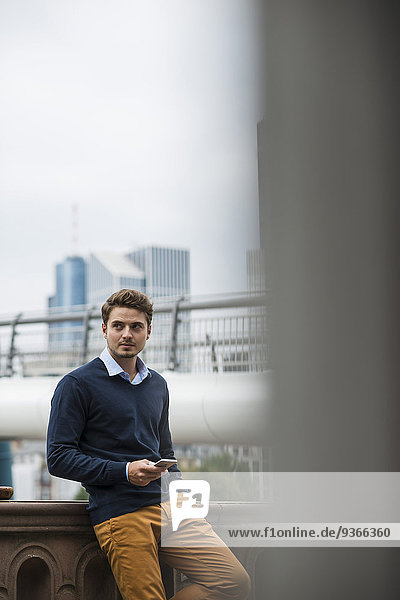 Deutschland  Hessen  Frankfurt  junger Mann auf einer Brücke stehend mit seinem Smartphone