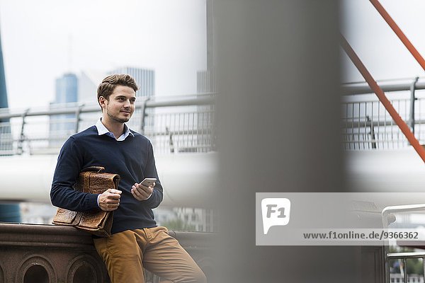 Deutschland  Hessen  Frankfurt  lächelnder junger Mann auf einer Brücke stehend mit Aktentasche und Smartphone