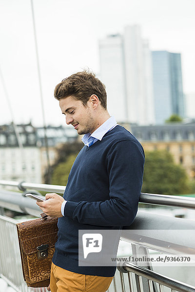 Deutschland  Hessen  Frankfurt  junger Geschäftsmann auf einer Brücke stehend mit seinem Smartphone
