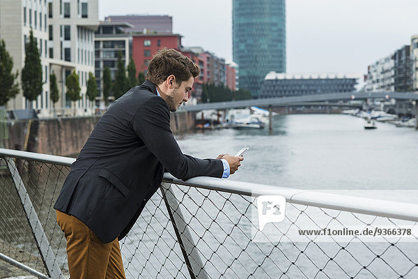 Deutschland  Hessen  Frankfurt  junger Geschäftsmann auf einer Brücke stehend mit seinem Smartphone