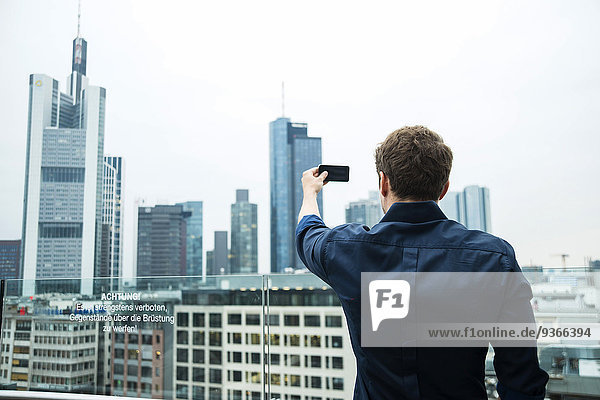 Deutschland  Hessen  Frankfurt  junger Mann beim Fotografieren der Skyline mit dem Smartphone