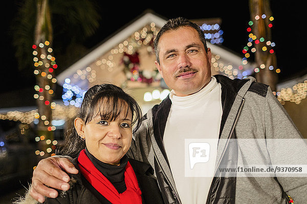 Hispanic couple smiling outside house decorated with string lights