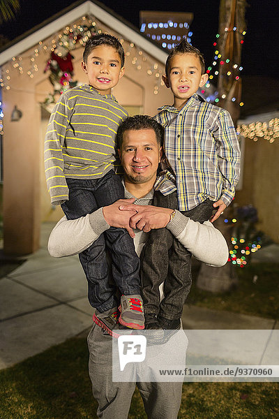 Hispanic man carrying sons on shoulders outside house decorated with string lights