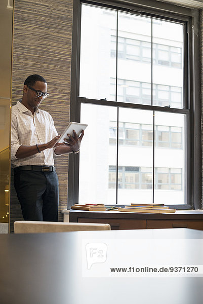 Ein Mann  der mit einem digitalen Tablett an einem Fenster in einem Büro steht.