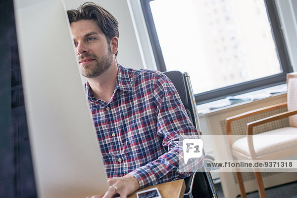Ein Mann in kariertem Hemd  der an einem Büroschreibtisch sitzt und einen Computer benutzt.
