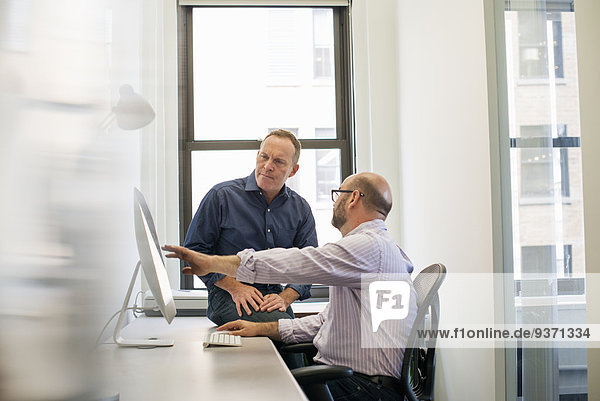 Zwei Geschäftskollegen in einem Büro  die sich unterhalten und sich auf einen Computerbildschirm beziehen.