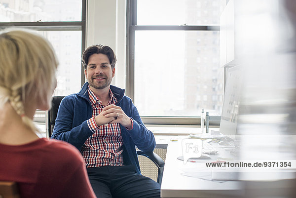 Office life. A man and woman in an office  seated talking to each other.