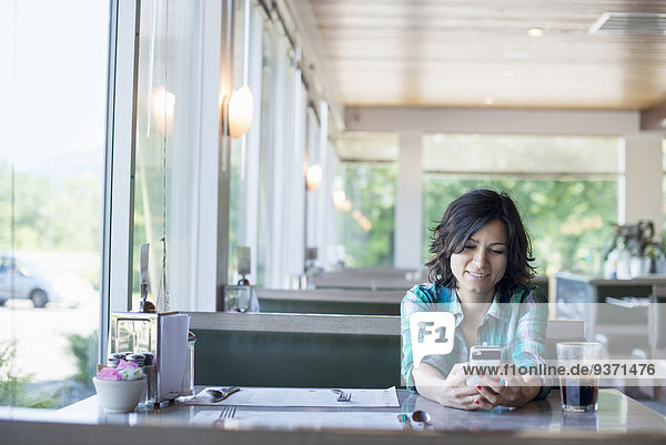 Eine Frau in kariertem Hemd  die lachend an einem Tisch sitzt und auf ihr Smartphone schaut.