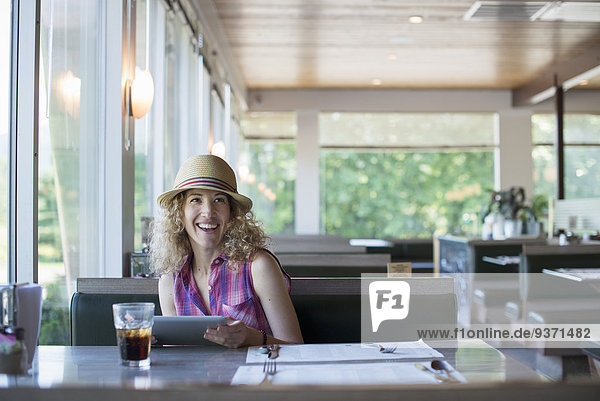 A woman in a hat sitting in a diner  holding a digital tablet.