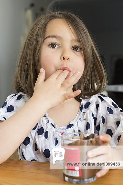 Kleines Mädchen isst Schokoladensauce aus einem Glas mit den Fingern