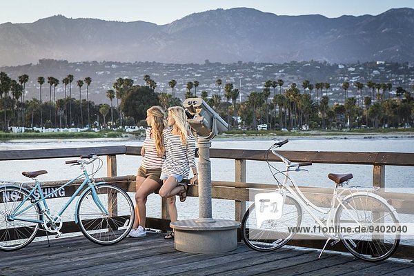 Two women in their twenties taking a break from riding their bikes. They are resting on the railing watching the sun go down with mountains and ocean in the background.