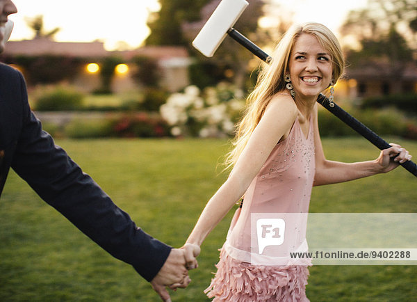 A beautiful young lady with croquet mallet on her left shoulder clutches the hand of her male partner.