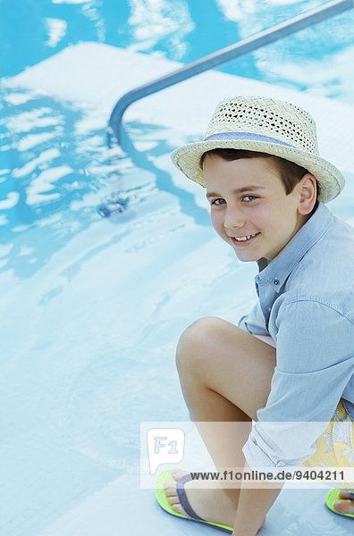 Porträt eines lächelnden Jungen mit Strohhut, der am Swimmingpool hockt