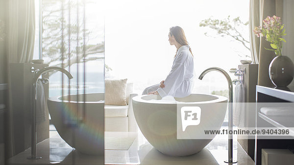Frau im weißen Bademantel  auf dem Wannenrand sitzend und durchs Fenster schauend