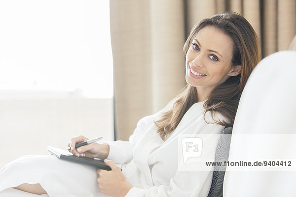 Portrait of smiling woman wearing white bathrobe with notebook in bedroom