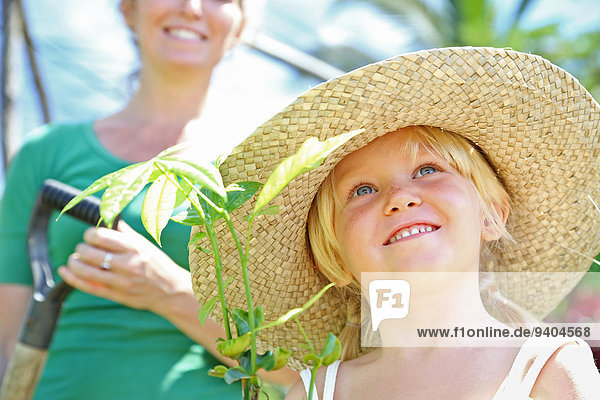 Mädchen mit Strohhut-Haltepflanze  Frau mit Schaufel im Hintergrund