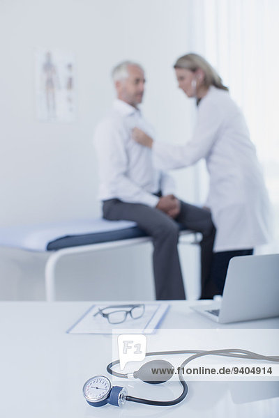 Blutdruckmessgerät  Laptop  Datei und Brille auf dem Schreibtisch in der Arztpraxis  Ärztin untersucht Patientin im Hintergrund