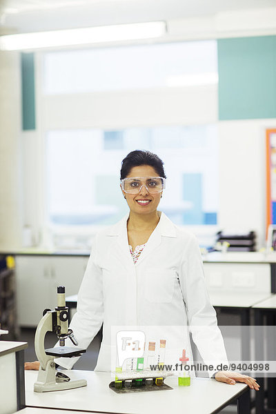 Porträt einer lächelnden Lehrerin mit Schutzbrille  die hinter dem Schreibtisch mit Mikroskop und Reagenzgläsern im Rack steht.