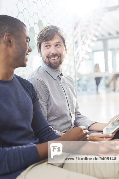 Zwei lächelnde Männer  die im Büroflur ein digitales Tablett halten.