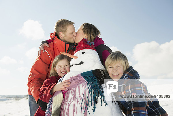 Girl and boy standing with snowman  parents kissing in background