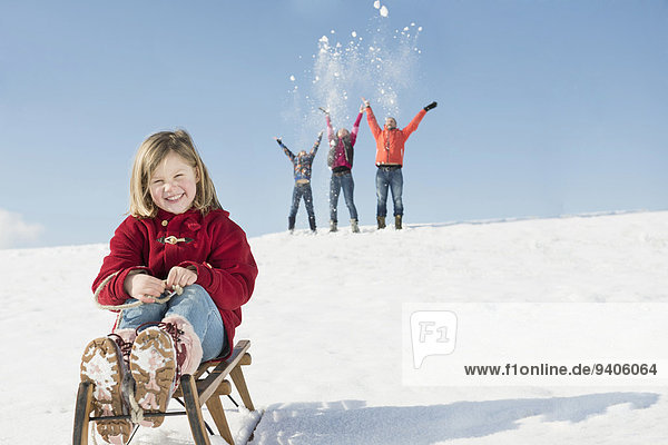 Portrait of girl sitting on sledge  family playing in background