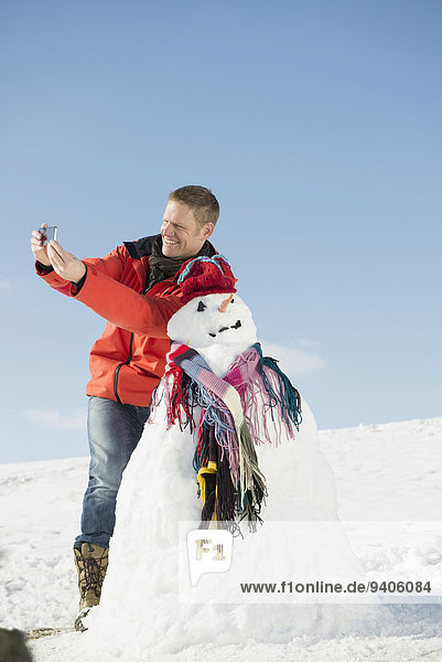 Man taking picture with smart phone of him and snowman