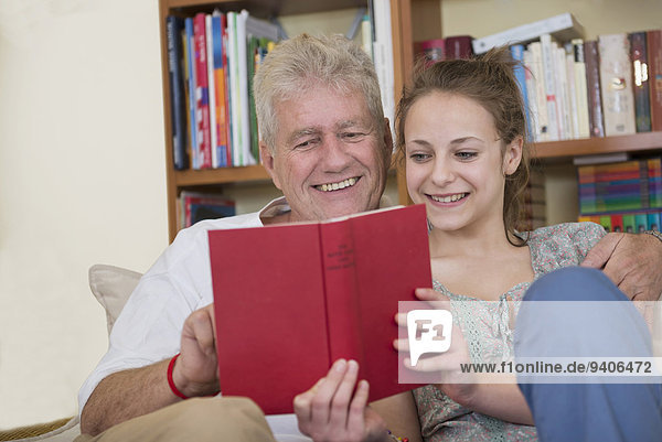 Grandfather and granddaughter reading book on couch in living room  smiling