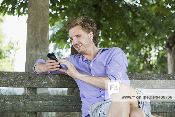 sitzend benutzen Mann lächeln Sitzbank Bank Mittelpunkt Smartphone Erwachsener