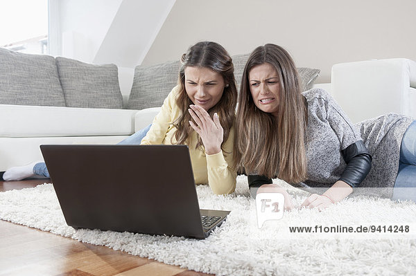 Two female friends lying side by side on carpet watching movie with laptop