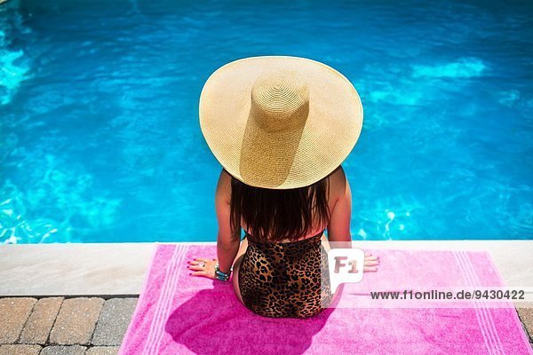 Rear view of young woman in sunhat sitting at poolside
