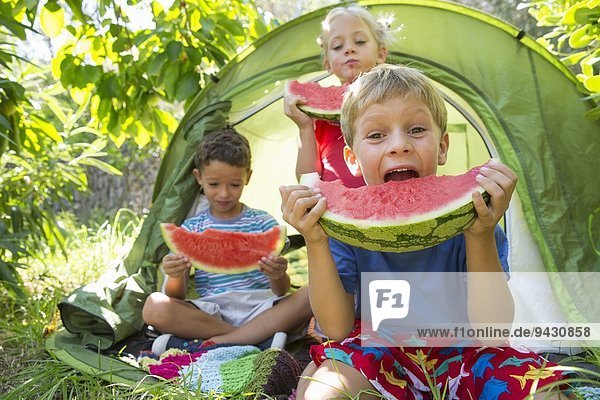 Drei Kinder essen große Wassermelonenscheiben im Gartenzelt