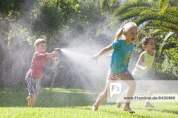 Drei Kinder im Garten jagen sich gegenseitig mit Wassersprenger