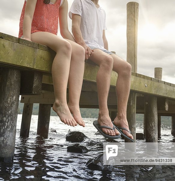 Junges Paar hält die Hände zusammen und sitzt am Steg über dem See.