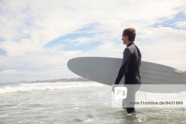 Junger Mann mit Surfbrett,  der aufs Meer hinausgeht.