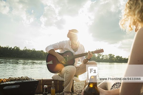 Young man sitting by lake playing guitar