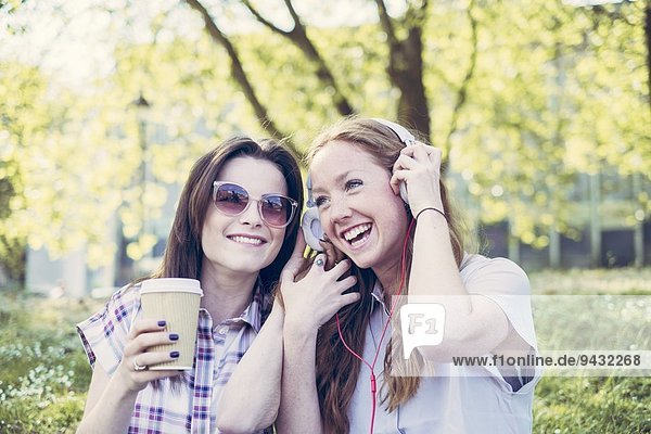 Zwei junge Freundinnen trinken Kaffee und hören Musik über Kopfhörer im Park.