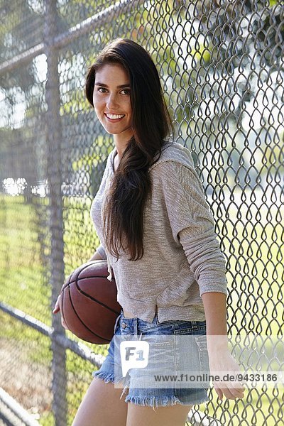 Young woman with basketball standing by wire fence