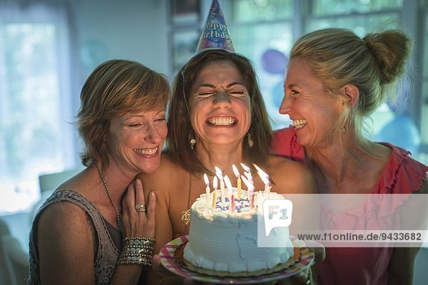 Reife Frau hält Geburtstagskuchen und wünscht sich,  während zwei Freunde zuschauen.