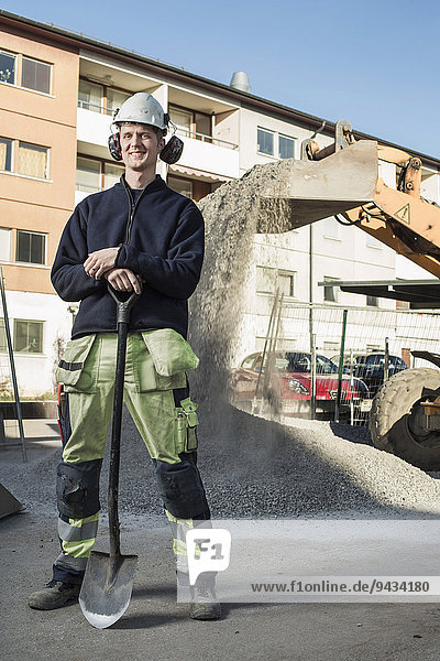 Ganzaufnahme eines lächelnden Bauarbeiters mit vor Ort stehender Schaufel