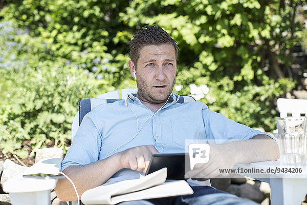 Mittlerer erwachsener Mann  der weg schaut  während er digitale Tabletten auf dem Sessel auf dem Hof benutzt.