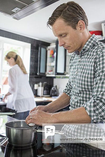 Ehemann beim Kochen  Frau beim Putzen von Utensilien im Hintergrund in der Küche