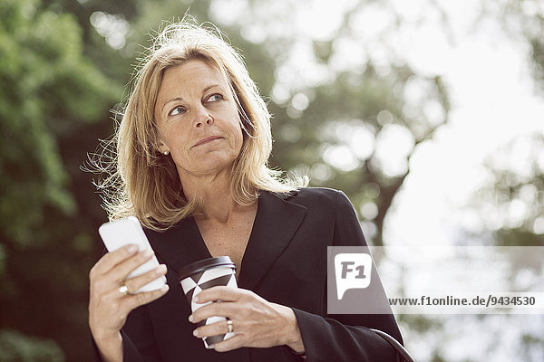 Geschäftsfrau schaut weg  während sie Smartphone und Einweg-Kaffeetasse im Freien hält.