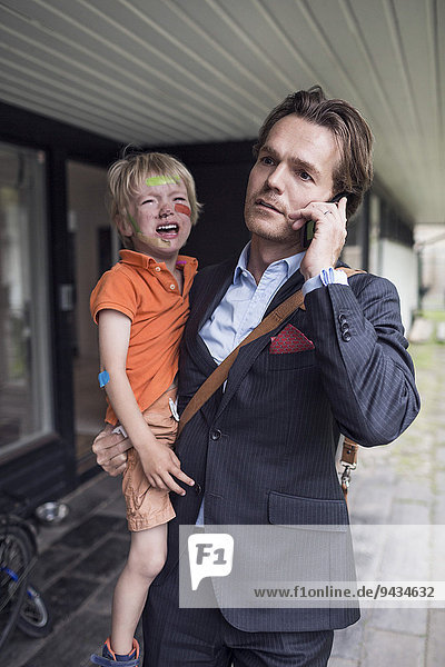 Geschäftsmann  der sein Handy benutzt  während er seinen weinenden Sohn aus dem Haus trägt.